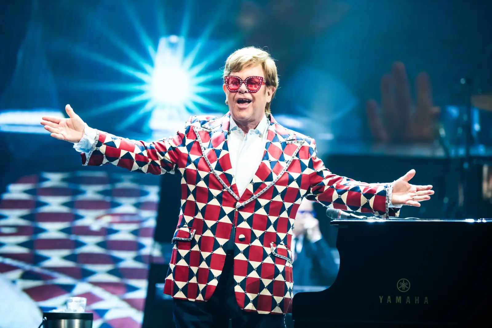 Elton John em apresentação ao vivo, de óculos vermelhos e braços abertos diante do piano
